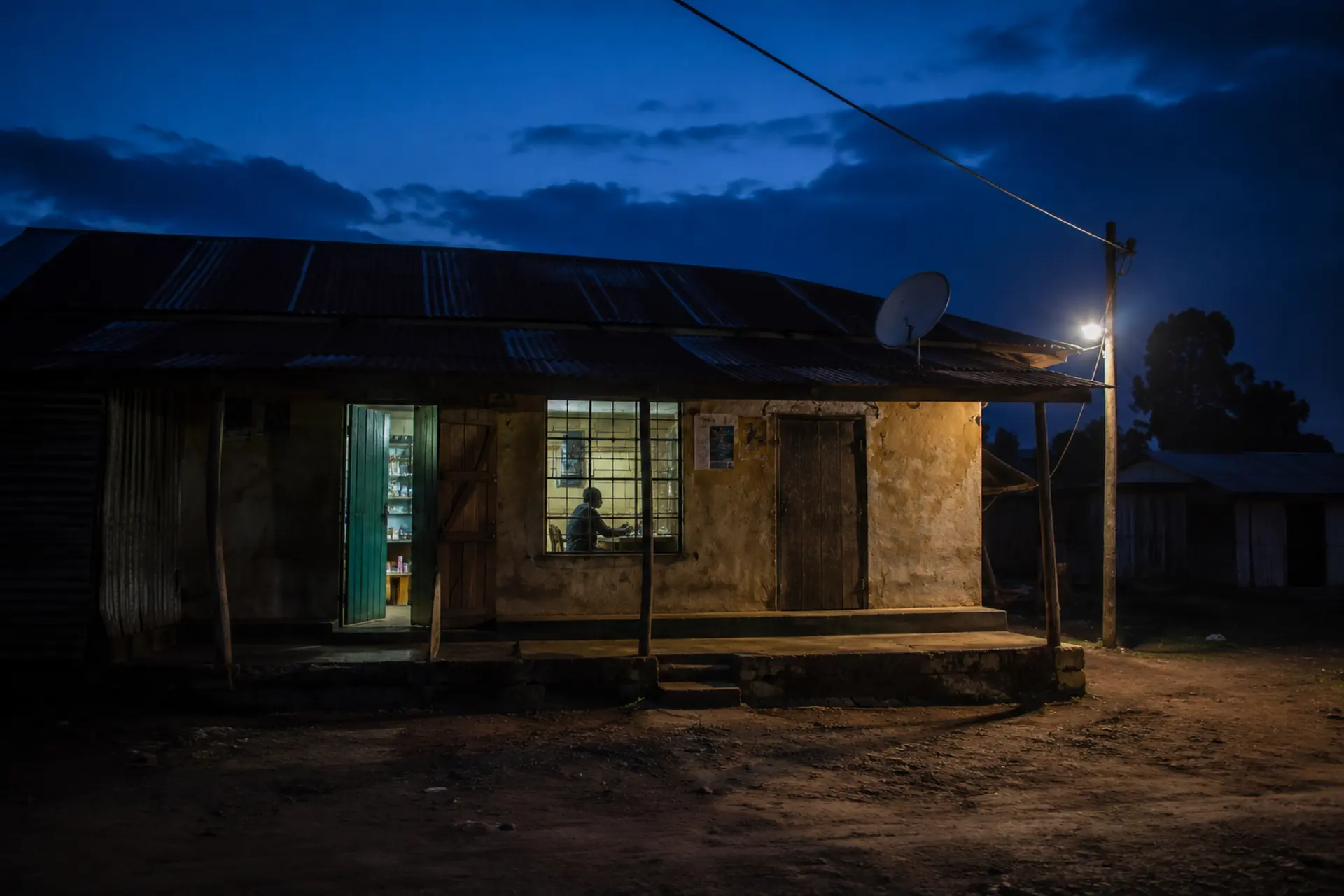A roadside duka at dusk, its interior lit and one person at the counter, with a security light and a satellite dish above the veranda.